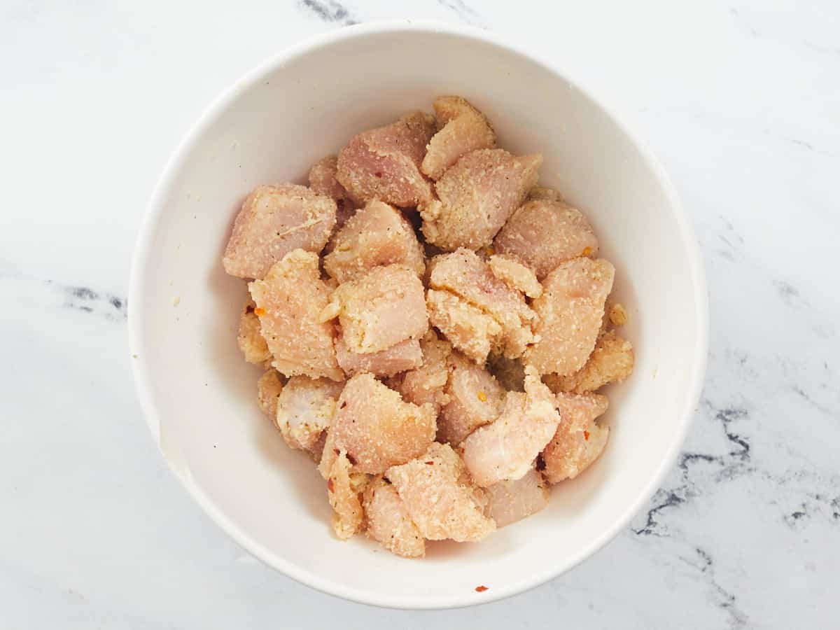 A white bowl of uncooked chicken breast chunks seasoned with grated Parmesan cheese, salt, black pepper, Italian seasoning and red pepper flakes. The bowl is sitting on a marble background. 