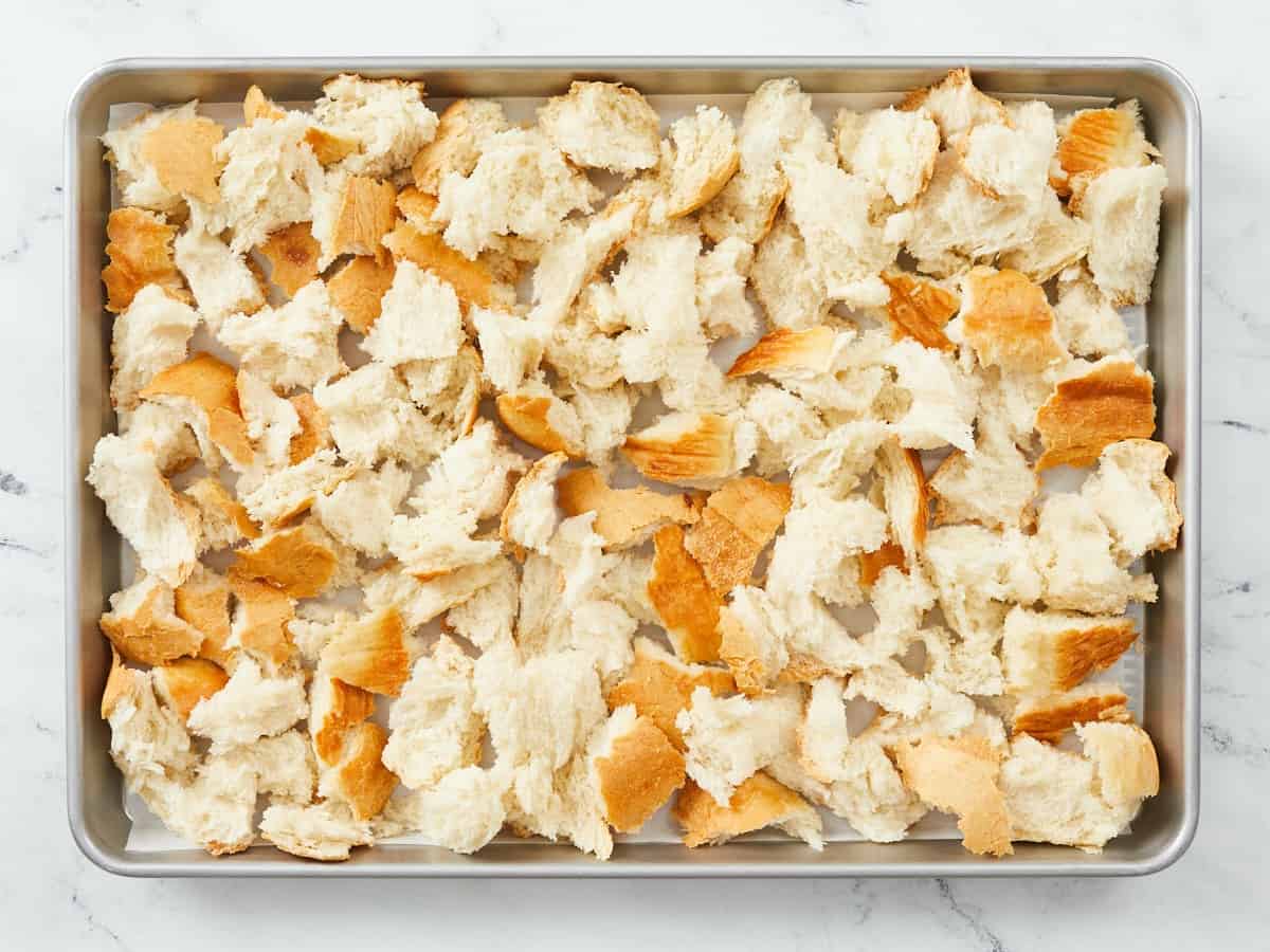 A rimmed metal baking sheet lined with parchment paper and covered with torn pieces of bread sitting  drying out to make a bread pudding, and the sheet pan is sitting on a white marble background.