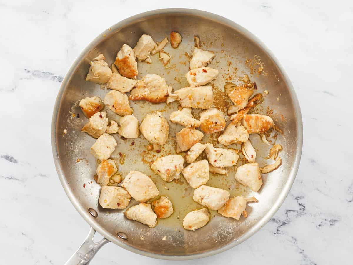 A large stainless steel skillet is sitting on a white marble background. In the skillet are chunks of cooked chicken breast and some olive oil.