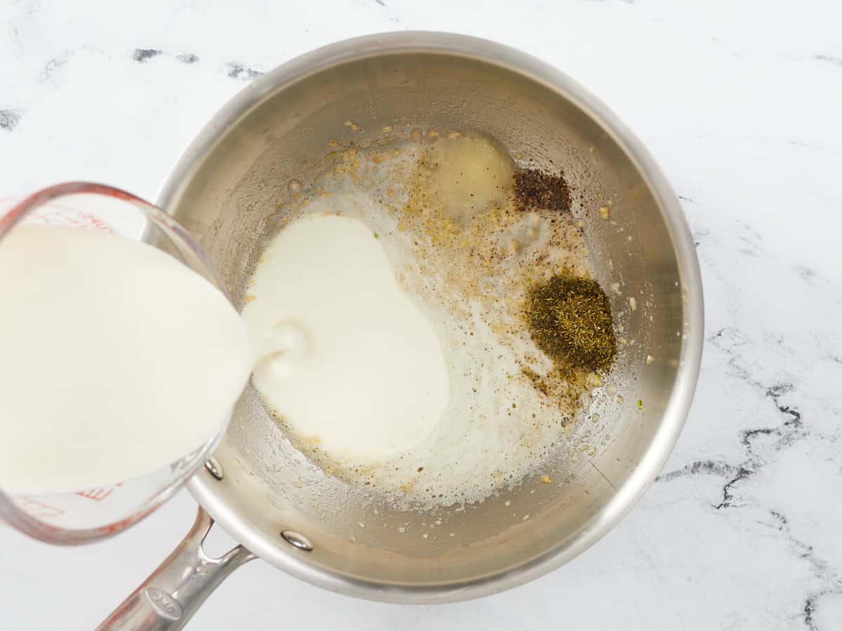 Heavy cream being poured into the left side of a stainless steel skillet with sauteed garlic, salt, pepper and italian season in seperate piles on the right side of the skillet.