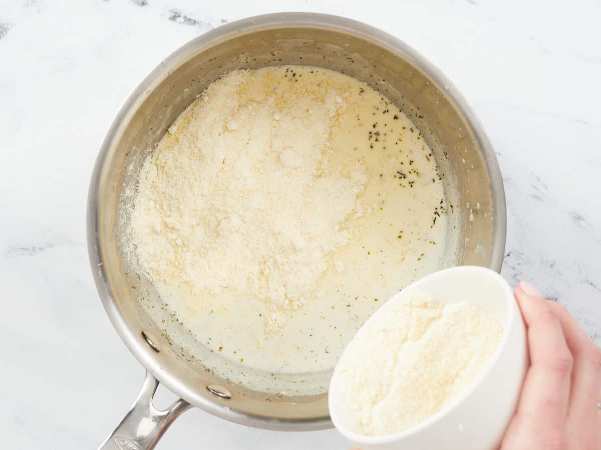 A hand dumping parmesan cheese from a small white bowl into the bottom right hand side of a stainless steel skillet that contains mixed heavy cream, melted butter, sauteed garlic and spices.