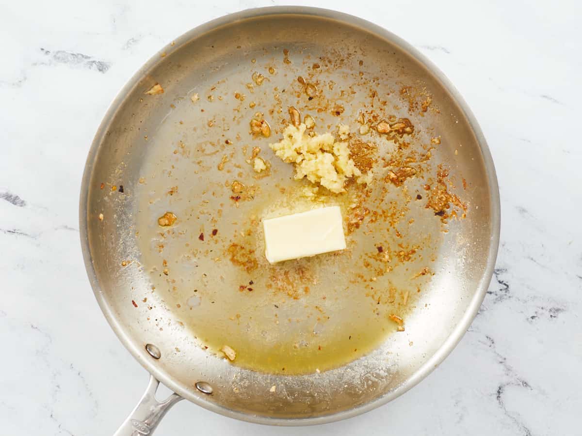A large stainless steel skillet on a background of white marble. The skillet is covered with fond after cooking chicken and there is a chunk of unmelted butter in the center and a pile of minced garlic. 