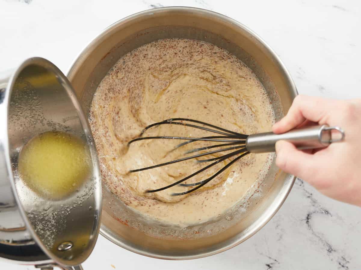 A large metal mixing bowl filled with the ingredients for bread pudding custard being whisked while a hand out of frame pours in melted butter from a small saucepan.