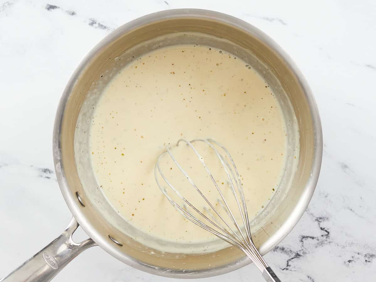 A whisk sitting in a stainless steel skillet after it was used to combine parmesan cheese with heavy cream, garlic, melted butter and spices to create alfredo sauce.