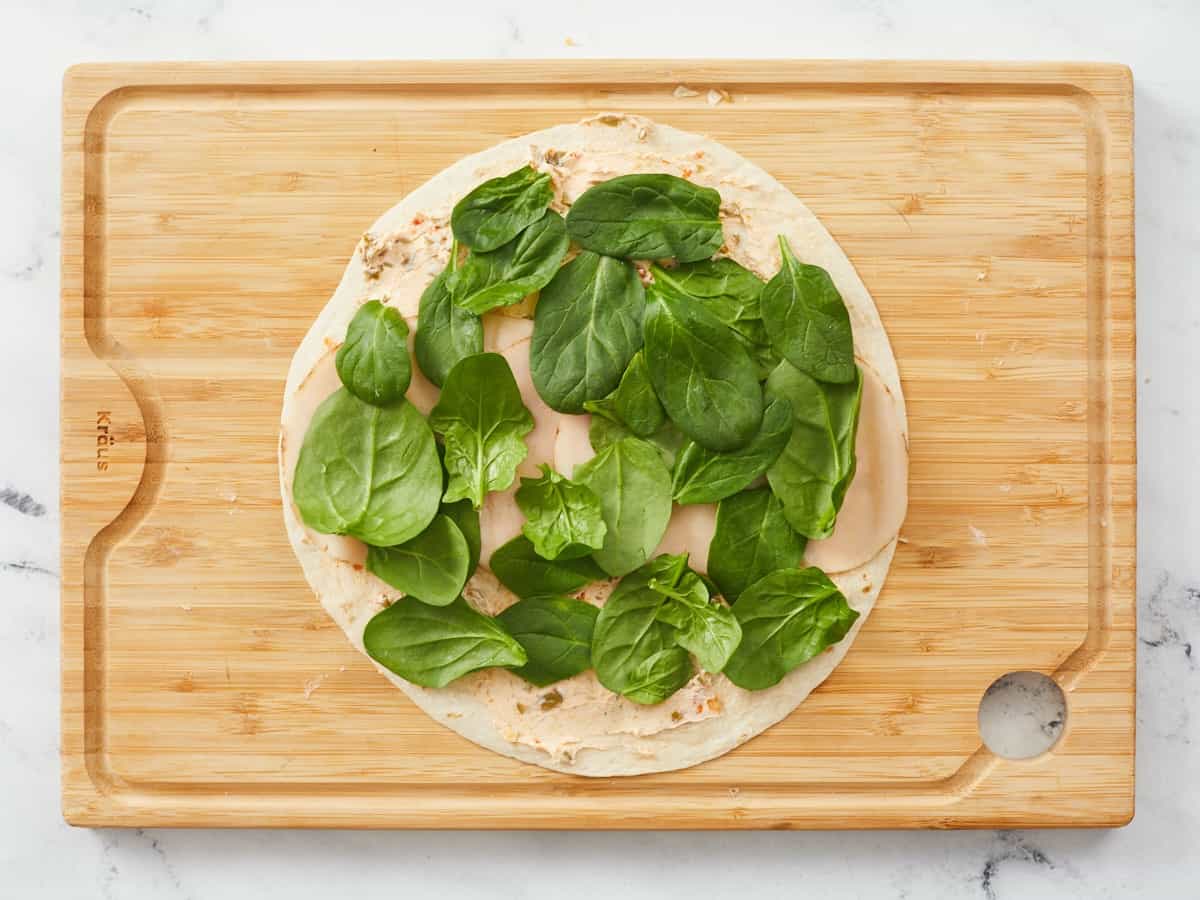 A large tortilla on a wooden cutting board covered with savory cream cheese, a row of turkey slices and covered in a single layer of baby spinach leaves. 
