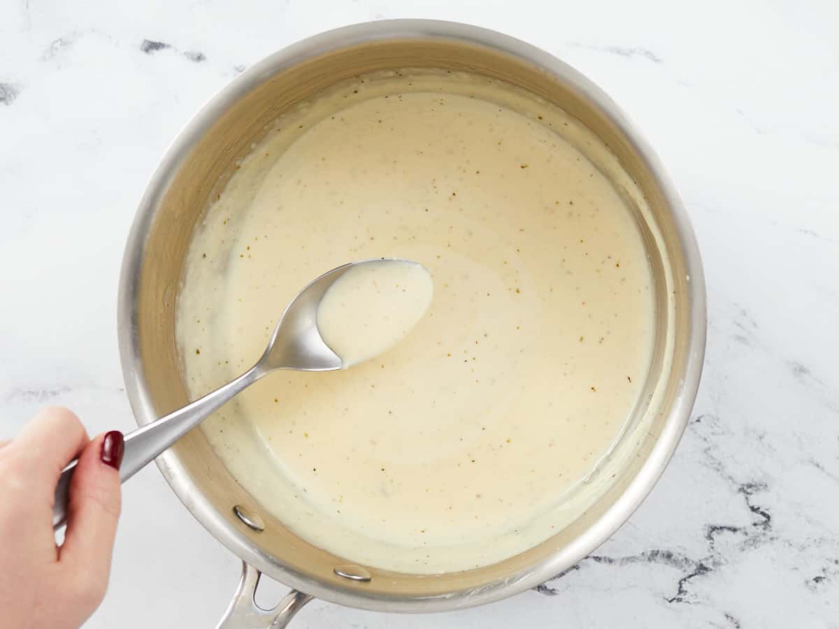 A hand is holding a metal serving spoon over a skillet of homemade alfredo sauce to show the thickness and consistency of the sauce once it has reduced and cooled slightly.