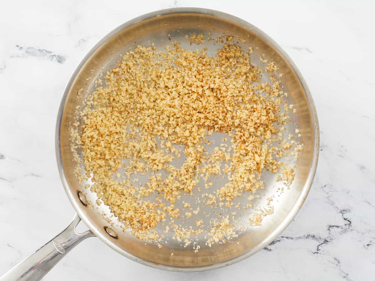 A stainless steel skillet filled with toasted, seasoned Panko breadcrumbs sitting on a white marble background.