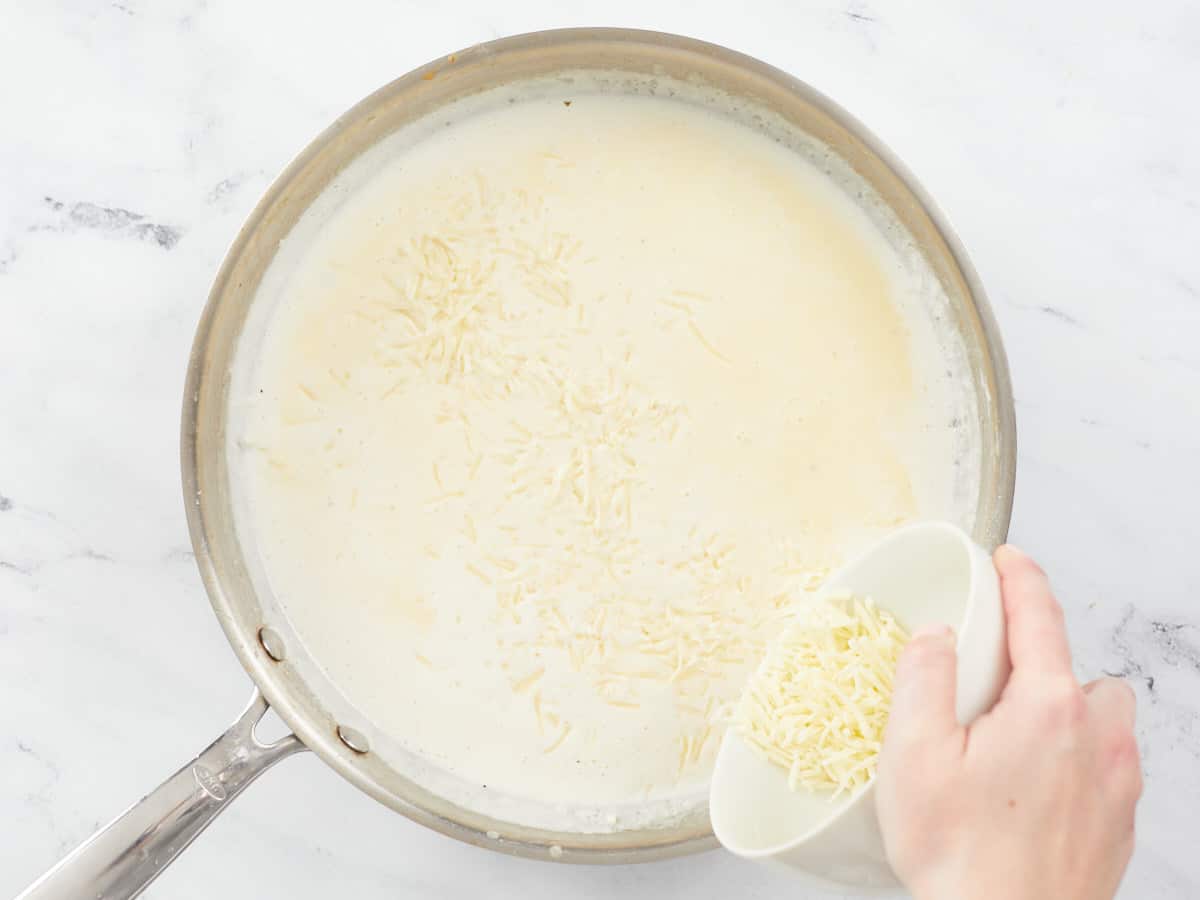 A large stainless steel skillet filled with heavy cream mixed with sautéed garlic, butter and seasonings. Grated parmesan cheese is sprinkled across the top of the heavy cream mixture from a small white bowl by a hand hovering over the bottom right side of the skillet.