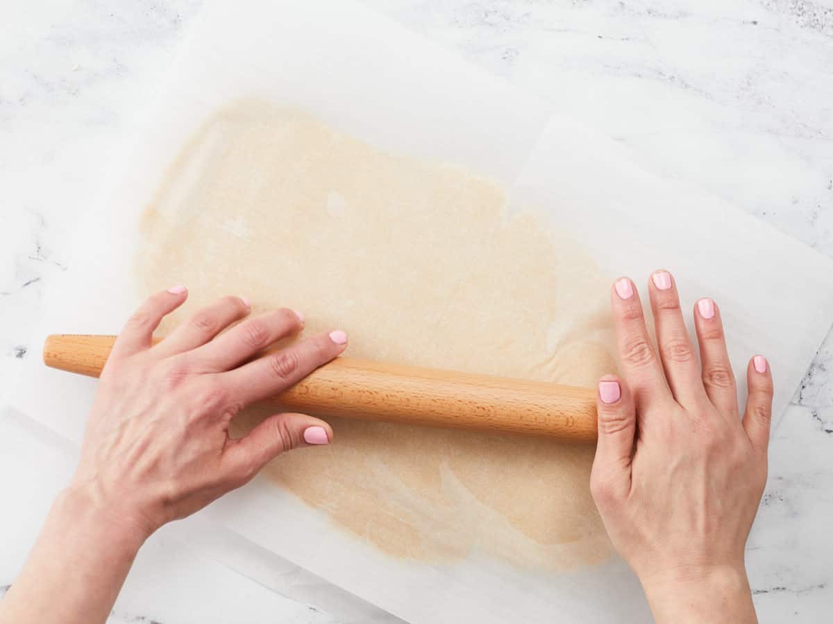 Two hands rolling out cookie dough between two sheets of parchment.