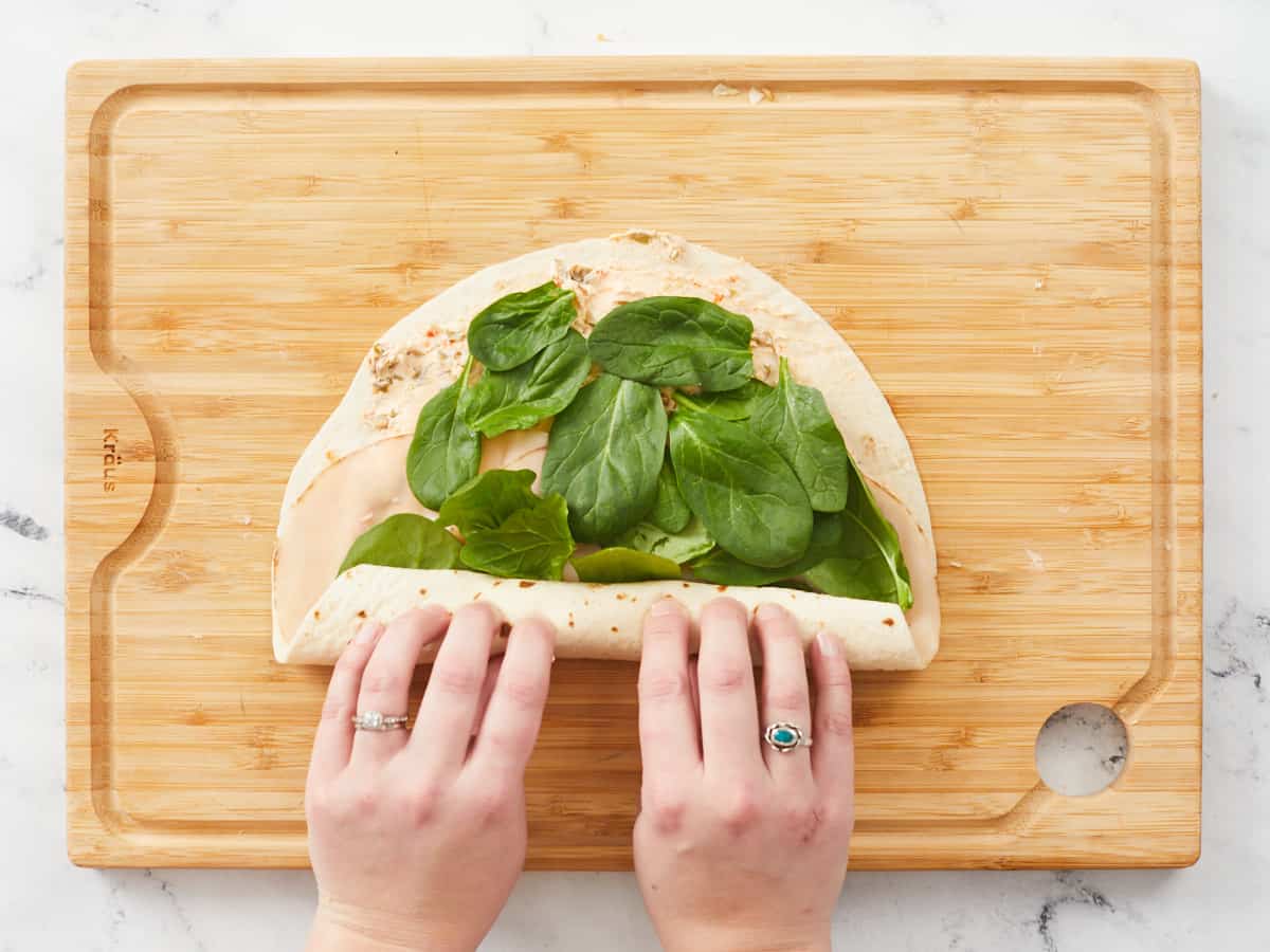 Hands rolling up a large tortilla on a wooden cutting board. The tortilla is covered with a savory cream cheese spread, a row of turkey slices and covered in a single layer of baby spinach leaves. 