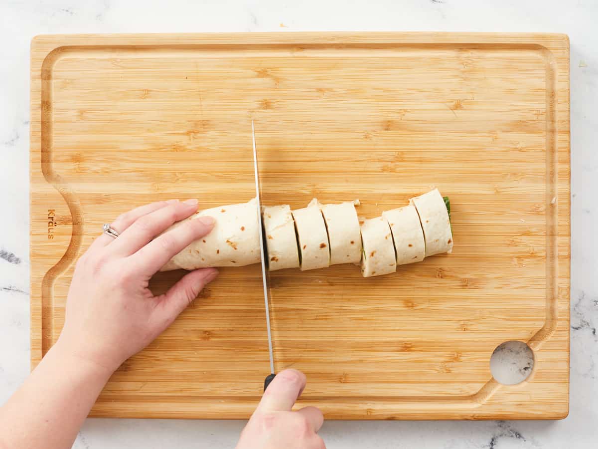 Two hands clicing a log of turkey pinwheels into one-inch pieces on a wooden cutting board.