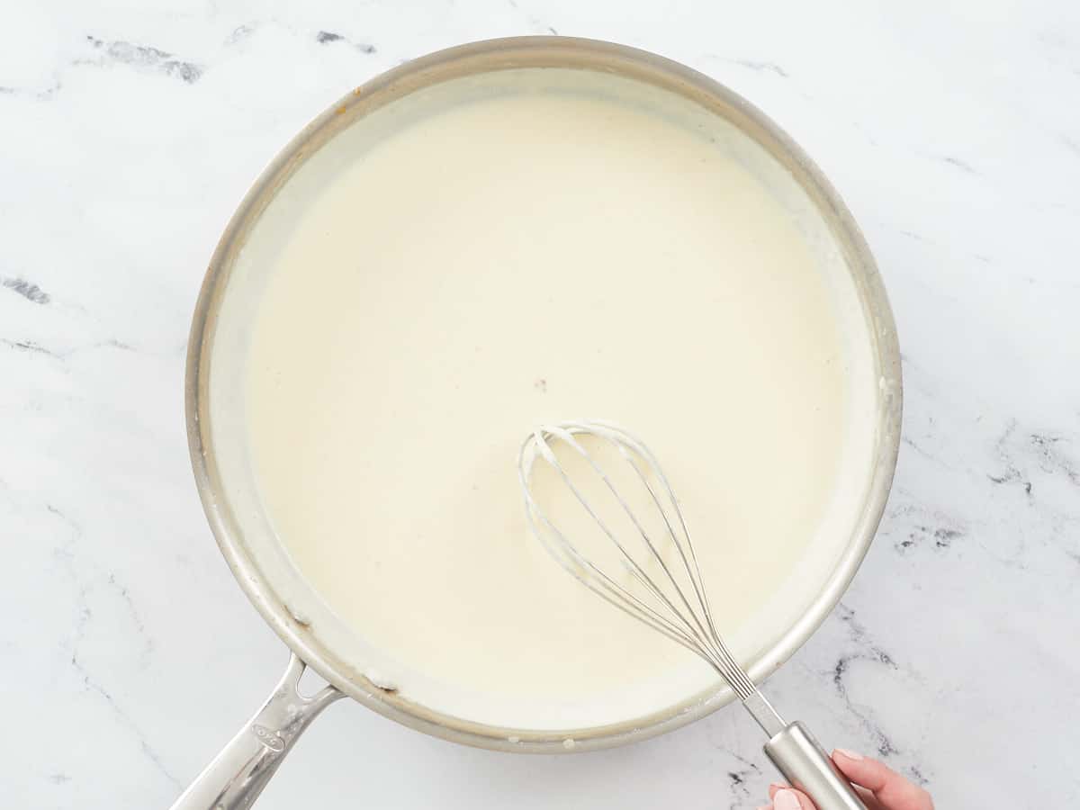 A large stainless steel skillet on a white marble background. The skillet is filled with a finished homemade alfredo sauce, and a hand in visible in the bottom right of the image and is stirring the mixture with a metal balloon whisk. 