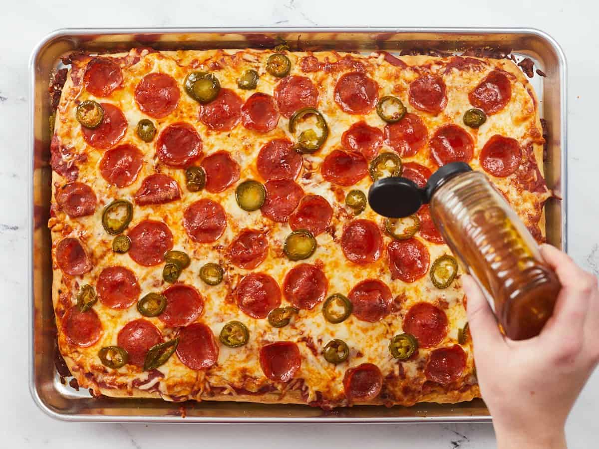 A fully baked sheet pan pizza that is golden brown and topped with crispy pepperonis, melted cheese and pickled jalapeño slices and a hand is drizzling honey on the right center of the pizza. 