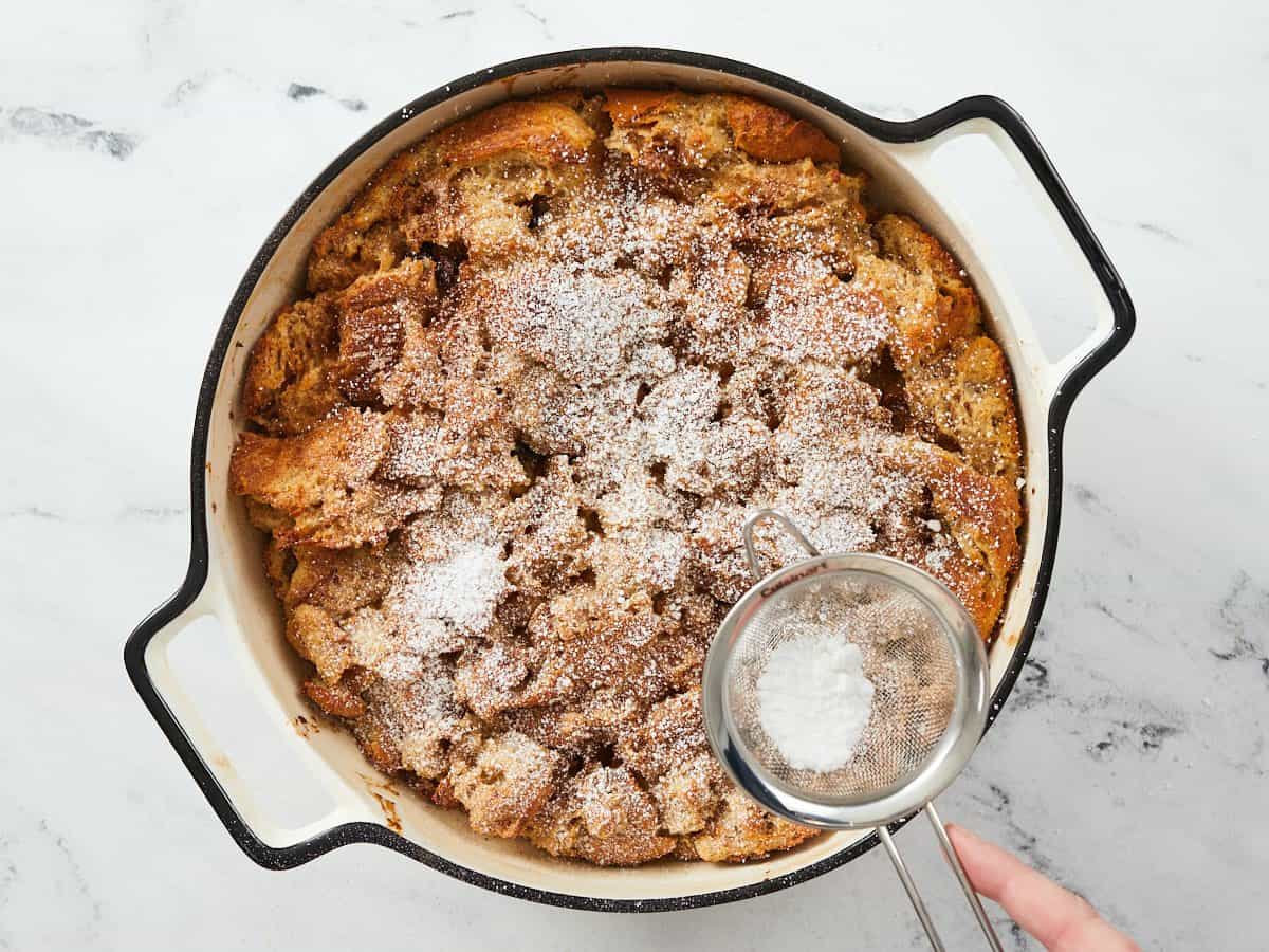A fully baked bread pudding in a round, white enamel baking dish being topped with powdered sugar in a small fine mesh strainer in the lower right hand side of the dish.