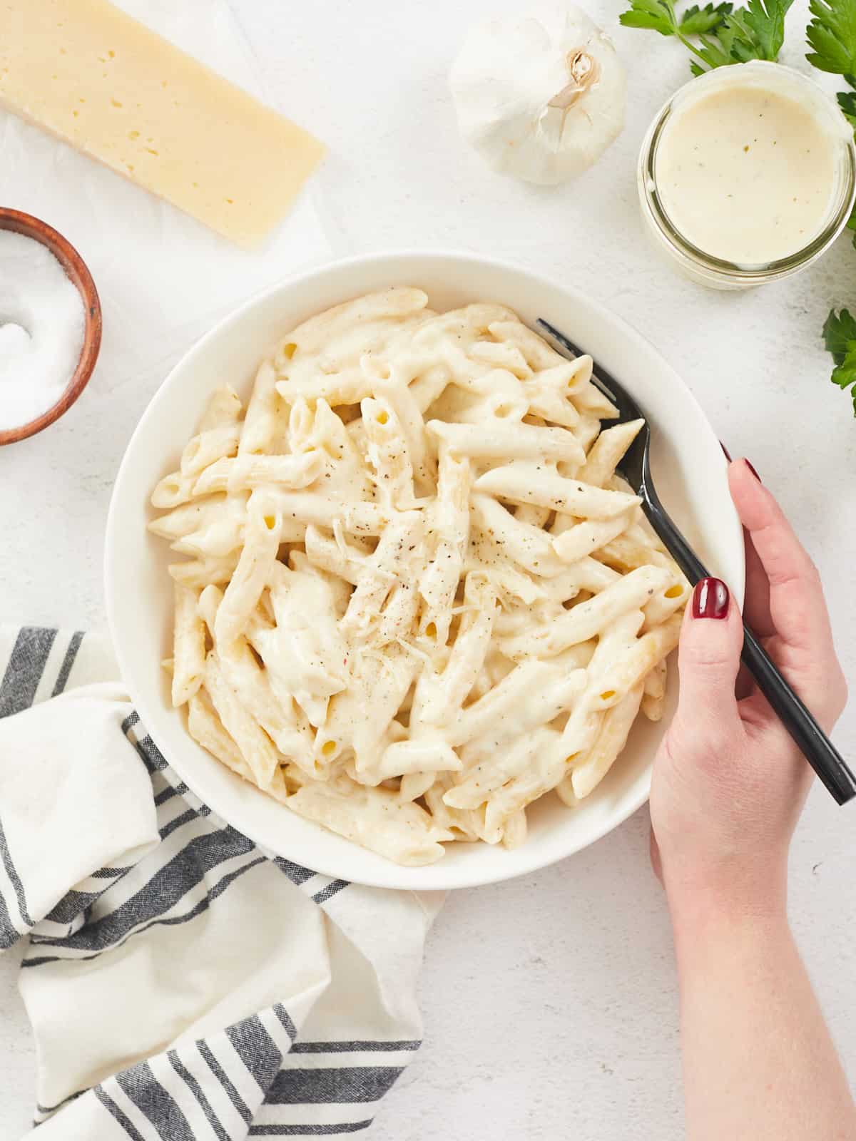 A large white bowl filled with cooked penne pasta dressed with homemade alfredo sauce, a hand is holding the right side of the bowl and a black metal fork, various props surround the bowl including a grey striped dish cloth, a block of parmesan cheese, a wooden bowl of salt and a mason jar of alfredo sauce.