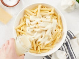 A large white bowl filled with cooked penne pasta, a hand is pouring alfredo sauce in the center of the bowl from a glass mason jar in the lower left side of the image, and there are various ingredients around the bowl mostly out of frame.