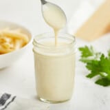A mason jar filled with alfredo sauce and a metal spoon raised above the jar after it's been dipped into the sauce so it's dripping off, and blurred in the background is a block of parmesean cheese, a bowl of uncooked pasta and some fresh italian parsley.