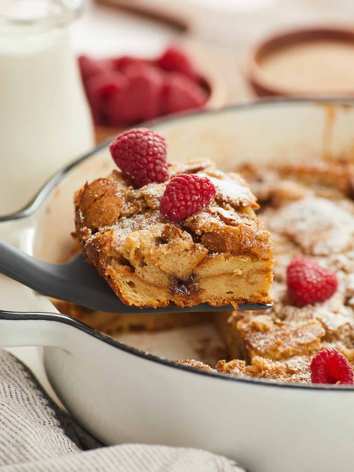 A vertical side view showing the cross section of slice of a baked bread pudding topped with powdered sugar and two raspberries as it is removed with a spatula in the left side of the frame, and blurred in the background are a small glass jar of heavy cream and wooden bowls filled with raspberries and cinnamon sugar..
