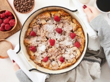 An overhead view of a baked bread pudding in round, white enamel cast iron baking dish topped with powdered sugar and fresh raspberries. There is a cutting board in the top left corner of the frame with a wooden bowl of raspberries and next to it is a bowl of chocolate chips and cup of coffee partially out of frame.