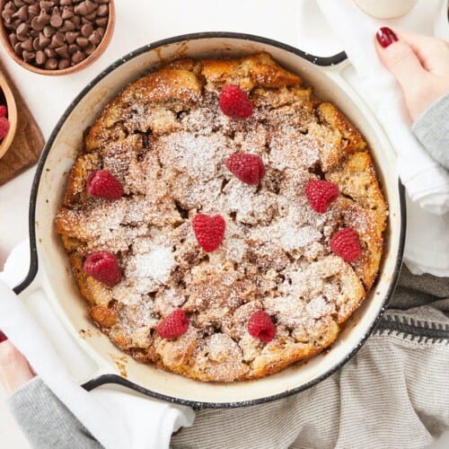 An overhead view of a baked bread pudding in round, white enamel cast iron baking dish topped with powdered sugar and fresh raspberries. There is a cutting board in the top left corner of the frame with a wooden bowl of raspberries and next to it is a bowl of chocolate chips and cup of coffee partially out of frame.