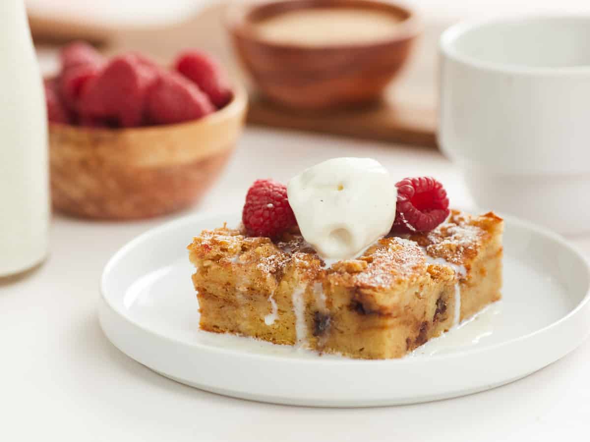 A horizontal image of a single serving of bread pudding on a flat white plate topped with two raspberries and a scoop of vanilla ice cream. In the background, there are blurred bowls of toppings ingredients in the recipe, like a small bottle of heavy cream, a bowl of fresh raspberries and a bowl of cinnamon sugar topping. 