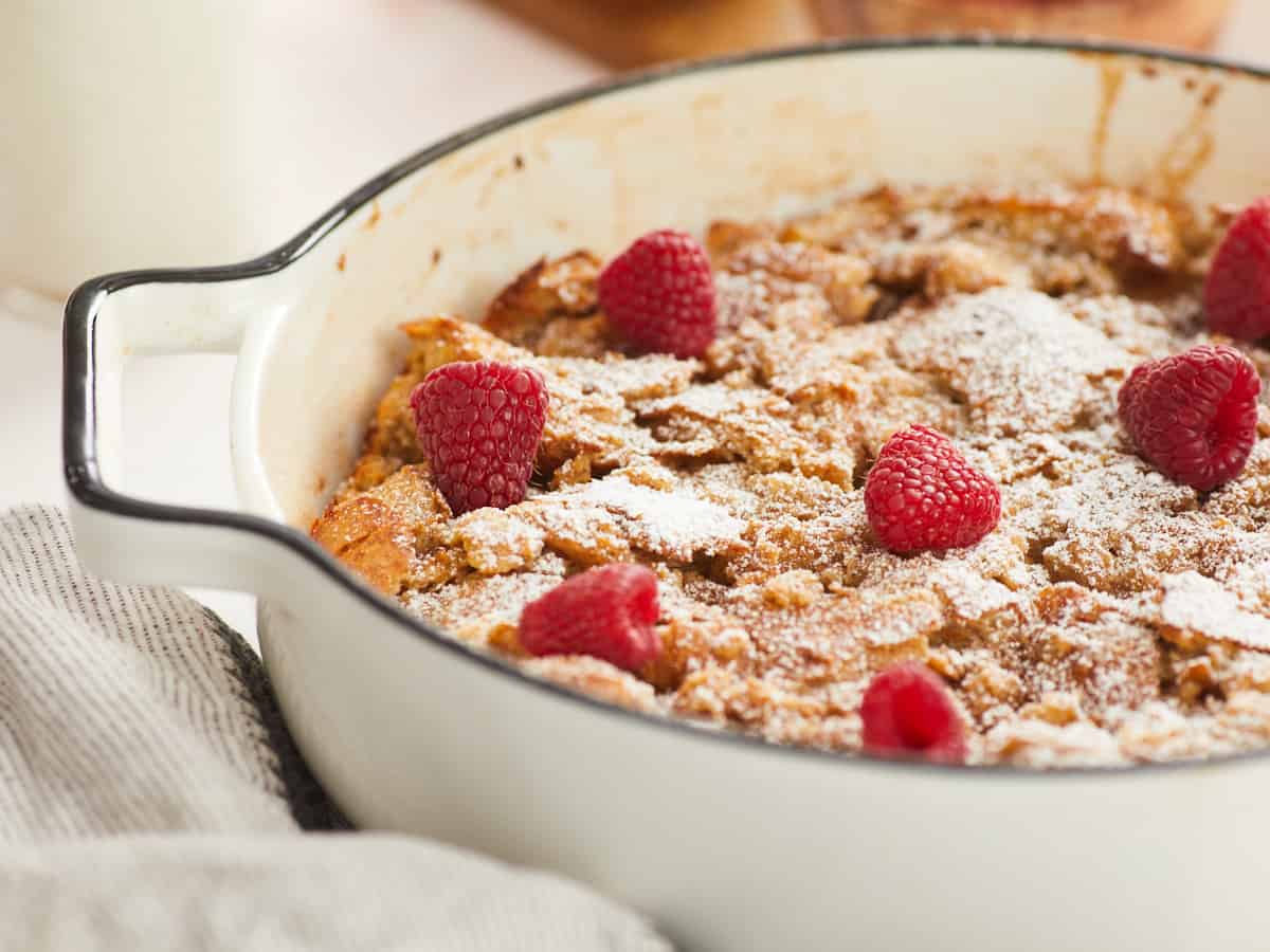 A close up, horizontal image displaying a side view of a baked bread pudding in a round, white enamel baking dish that is topped with powdered sugar and fresh raspberries.