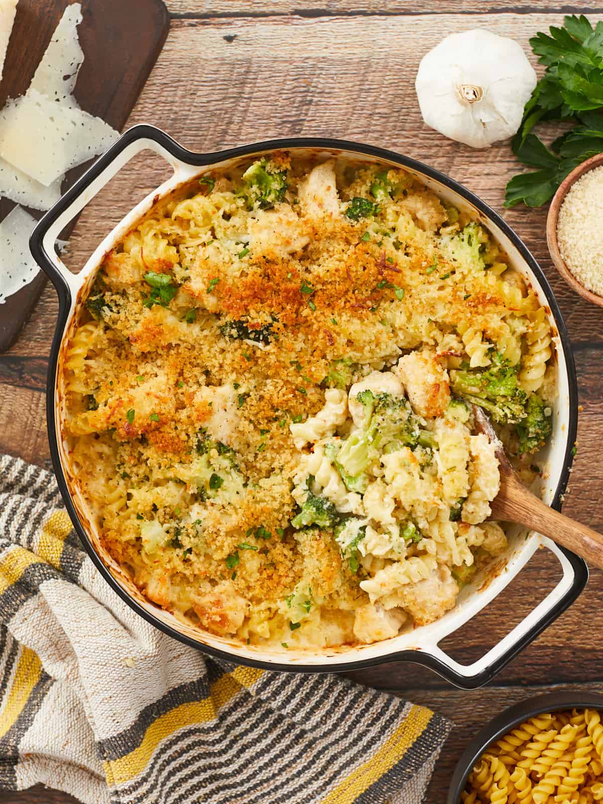 A golden brown chicken alfredo bake in a round, white enamel casserole dish on a wooden background surrounded by a yellow and gray striped dish towel in the bottom left, a wooden cutting board with shaved Parmesan cheese in the top left corner, a clove of garlic, sprigs of parsley, and small wooden bowl of breadcrumbs in the top right corner, and a small wooden bowl of uncooked pasta in the bottom right corner. The right side of the casserole has been stirred with a large wooden spoon that's resting in the dish. 