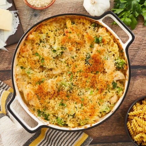 A golden brown chicken alfredo bake in a round, white enamel casserole dish on a wooden background surrounded by a yellow and gray striped dish towel in the bottom left, a wooden cutting board with shaved Parmesan cheese in the top left corner, a clove of garlic, sprigs of parsley, and small wooden bowl of breadcrumbs in the top right corner, and a small wooden bowl of uncooked pasta in the bottom right corner.