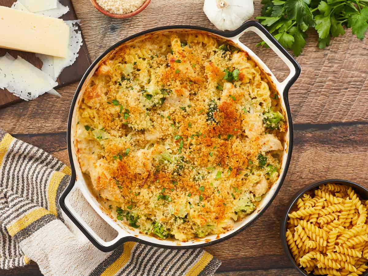 A golden brown chicken alfredo bake in a round, white enamel casserole dish on a wooden background surrounded by a yellow and gray striped dish towel in the bottom left, a wooden cutting board with shaved Parmesan cheese in the top left corner, a clove of garlic, sprigs of parsley, and small wooden bowl of breadcrumbs in the top right corner, and a small wooden bowl of uncooked pasta in the bottom right corner.