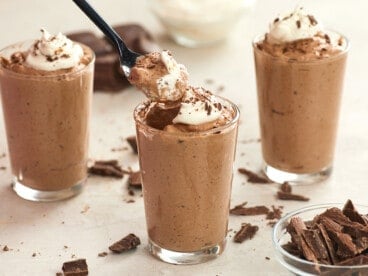 A spoon digging into a cup of chocolate mousse with whipped cream.