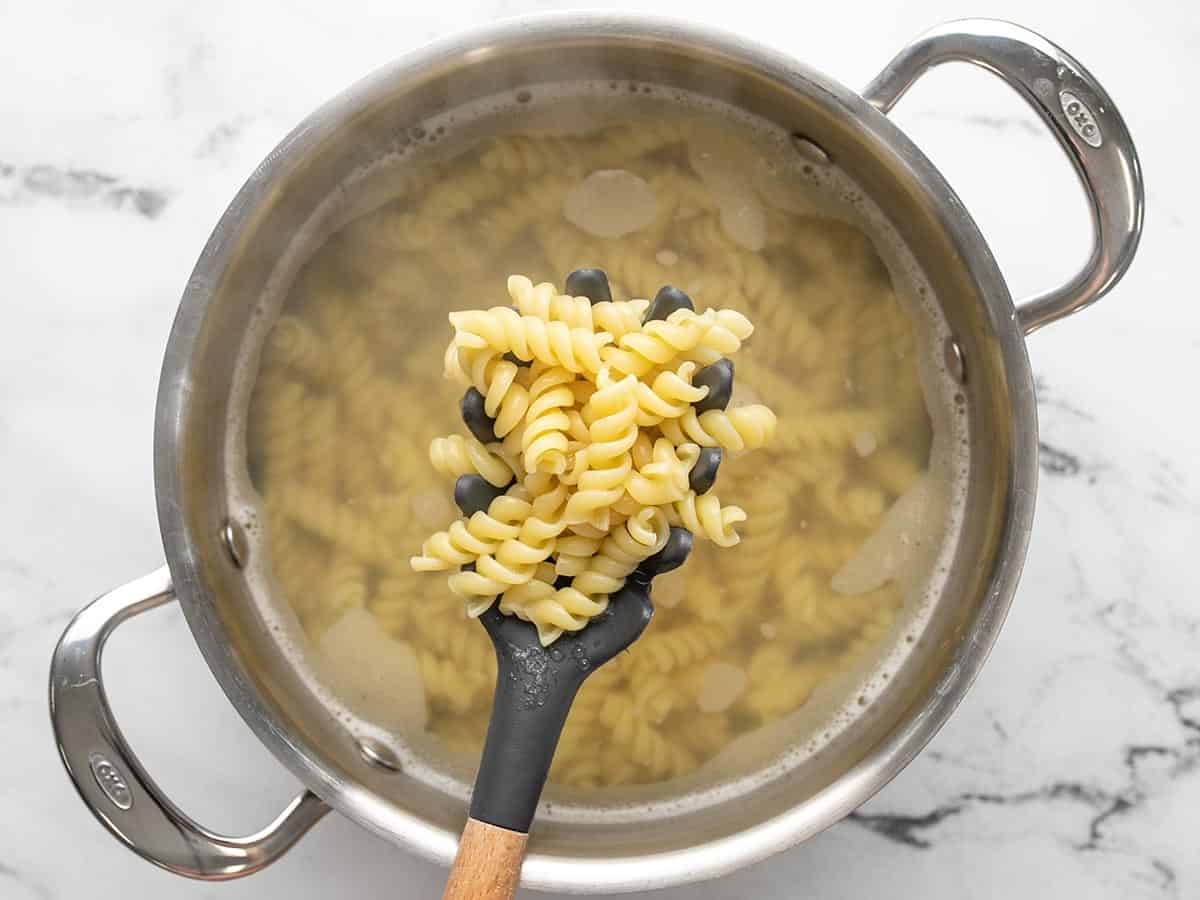 A medium-size stainless steel pot filled with cloudy pasta water and cooked rotini. A wooden spoon with a gray, silicon spoon is being held above the pot to show that the pasta is fully cooked. The pot is sitting on a white marble backdrop.
