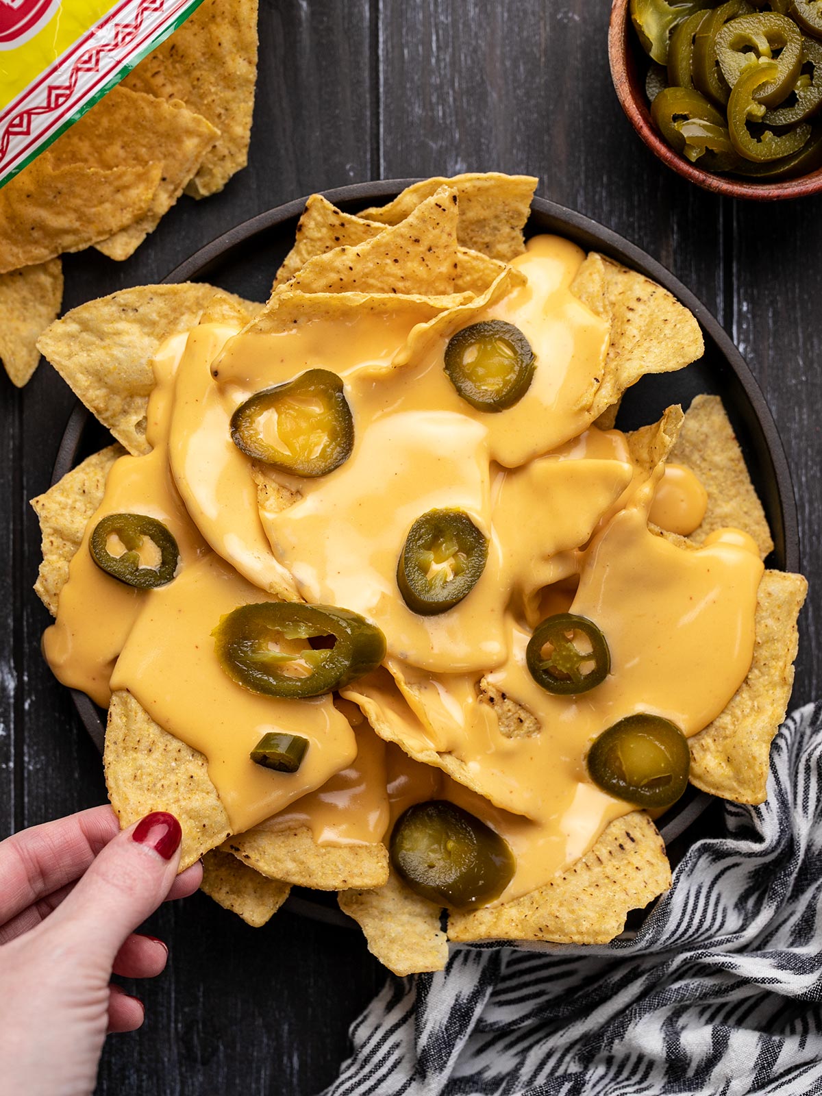 A plate of nachos with pickled jalapeño and a hand taking one chip.