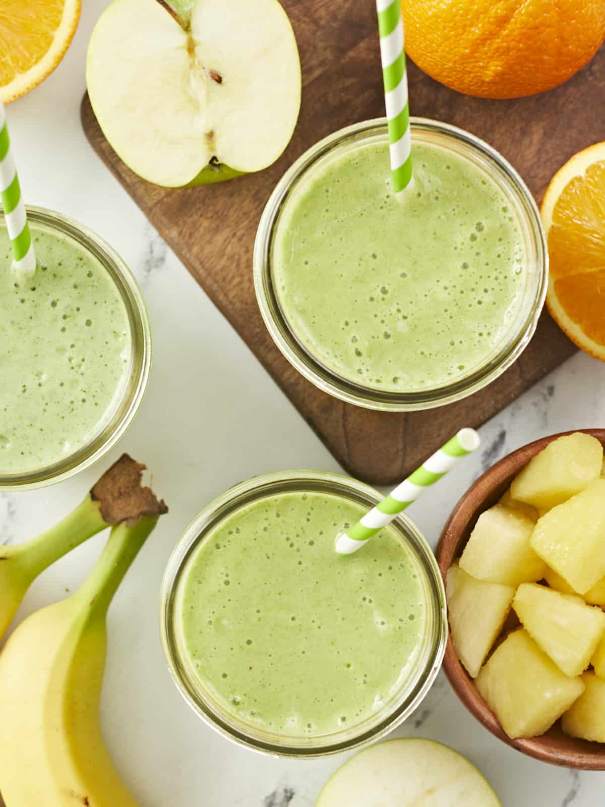 Overhead shot of three green smoothies surrounded by cut fruit.