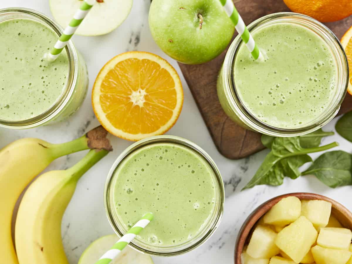 Overhead shot of three green smoothies surrounded by cut fruit.