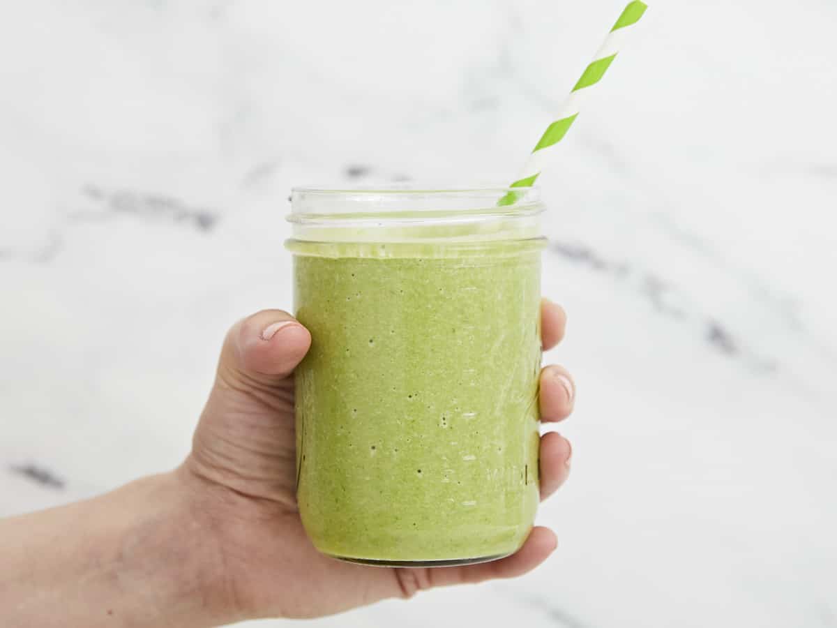 Side shot of hand holding green smoothie with a straw in it.