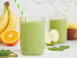 Side shot of green smoothies surrounded by cut fruit.