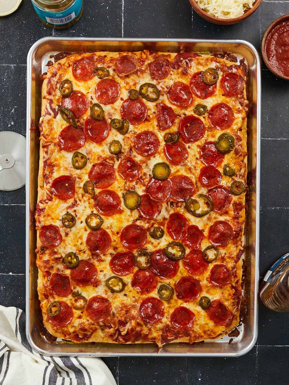 A vertical image of a baked sheet pan pizza topped with pizza sauce, cheese, pepperonis, pickled jalepenos and honey that is not sliced.