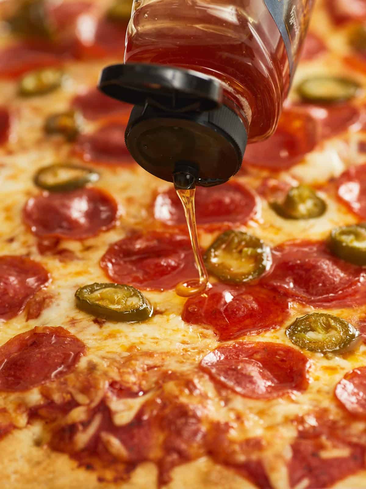 An extreme close up of baked sheet pan pizza topped with pepperonis and jalepenos that's being drizzled with honey.