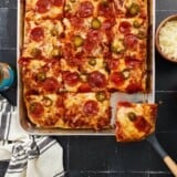 A vertical image of a baked sheet pan pizza topped with pizza sauce, cheese, pepperonis, pickled jalepenos and honey sliced and a spatula is removing the bottom right slice.