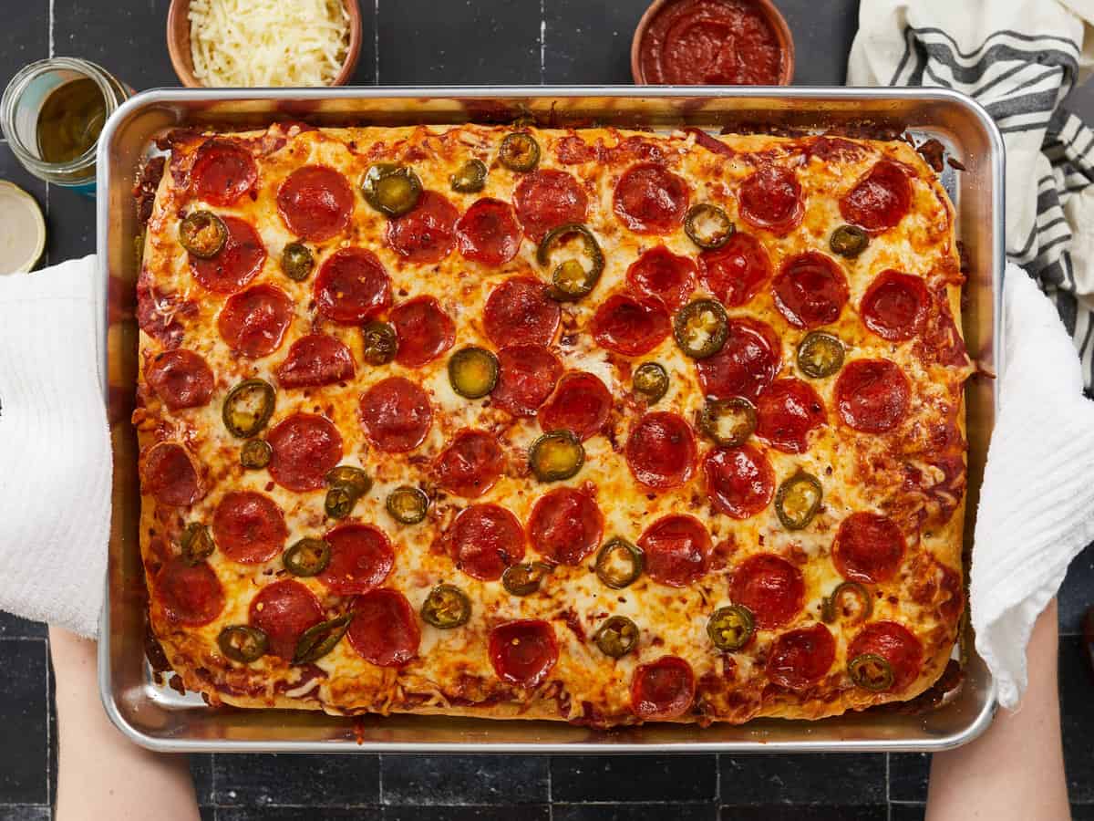 A horizontal image of a fully baked sheet pan pizza topped with pizza sauce, shredded cheese, pepperonis, jalepenos and honey that's held up with two white dish towels.