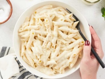 A large white bowl filled with cooked penne pasta dressed with homemade alfredo sauce, a hand is holding the right side of the bowl and a black metal fork, various props surround the bowl including a grey striped dish cloth, a block of parmesan cheese, a wooden bowl of salt and a mason jar of alfredo sauce.