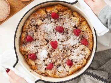 A vertical image of a baked bread pudding in round, white enamel cast iron baking dish topped with powdered sugar and fresh raspberries. There is a cutting board in the top left corner of the frame with a wooden bowl of raspberries and next to it is a cup of coffee partially out of frame.