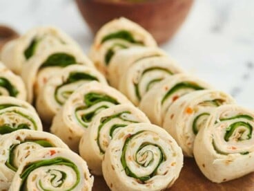 A close up shot of Turkey Pinwheels arranged in three rows on a wooden cutting board with a white background and a bowl of spinach blurred in the background