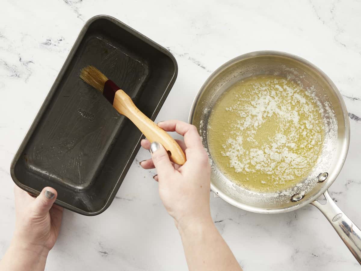An overhead shot of two hands preparing a metal loaf pan with a pastry brush next to a small skillet of melted butter.