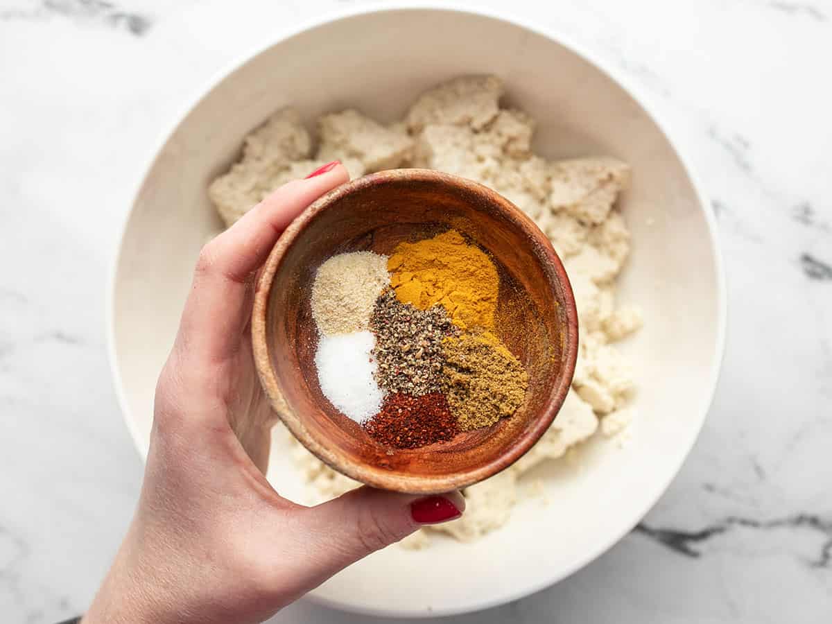 Spices in a bowl, unmixed, held over the bowl of tofu.