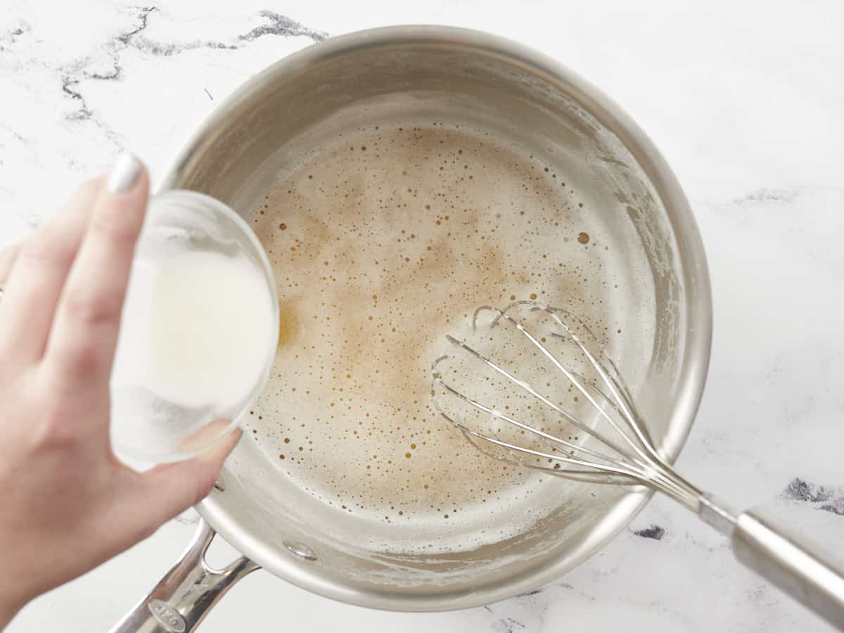 A cornstarch slurry is being poured into the left side of a small saucepot of beer, and a whisk is resting in the bottom right corner of the pot. 