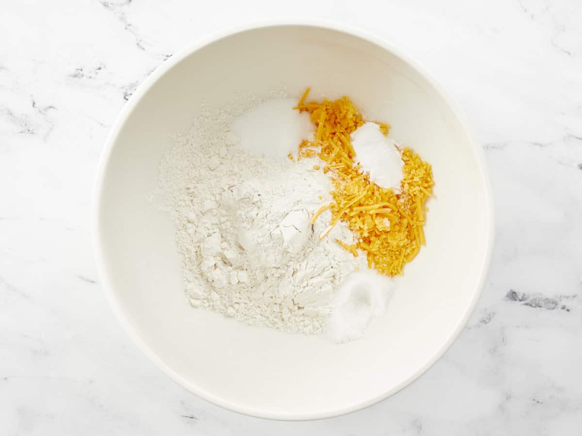 A white ceramic mixing bowl filled with piles of unmixed flour, sugar, baking powder, salt, sugar and shredded cheddar cheese.