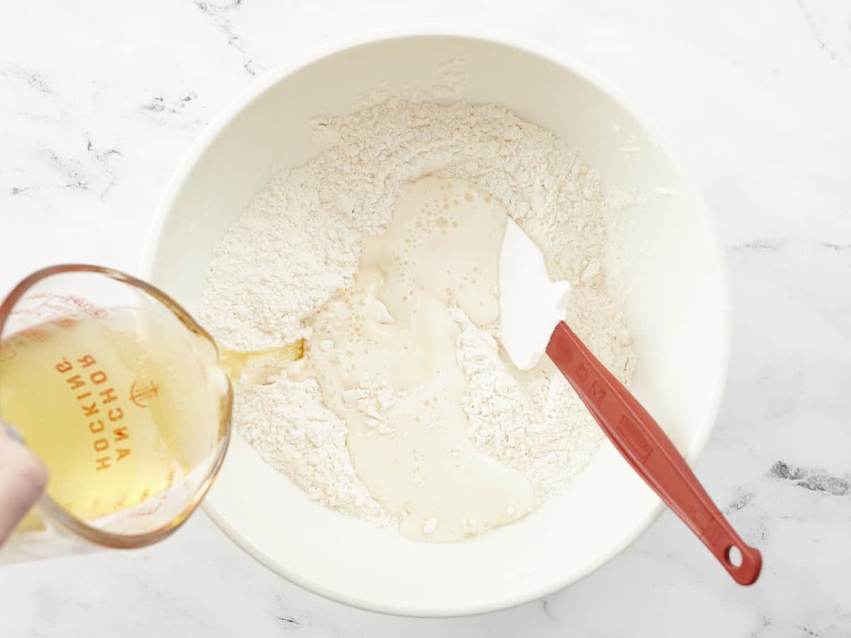 A white, ceramic mixing bowl filled with the mixed dry ingredients to make beer bread, a spatula is resting in the right side of the bowl and beer is being poured into the left side of the bowl from a glass liquid measuring cup.