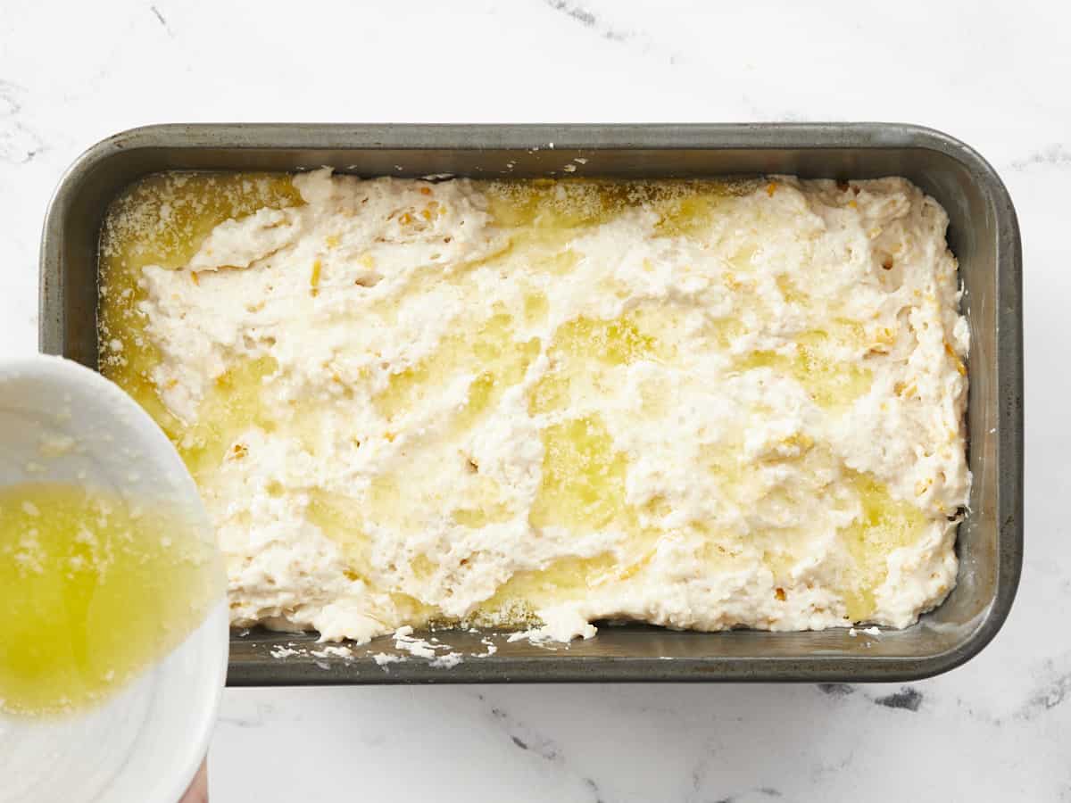 A metal loaf pan filled with unbaked bread dough is being covered with melted butter poured from a small, white bowl on the left side of the frame. 