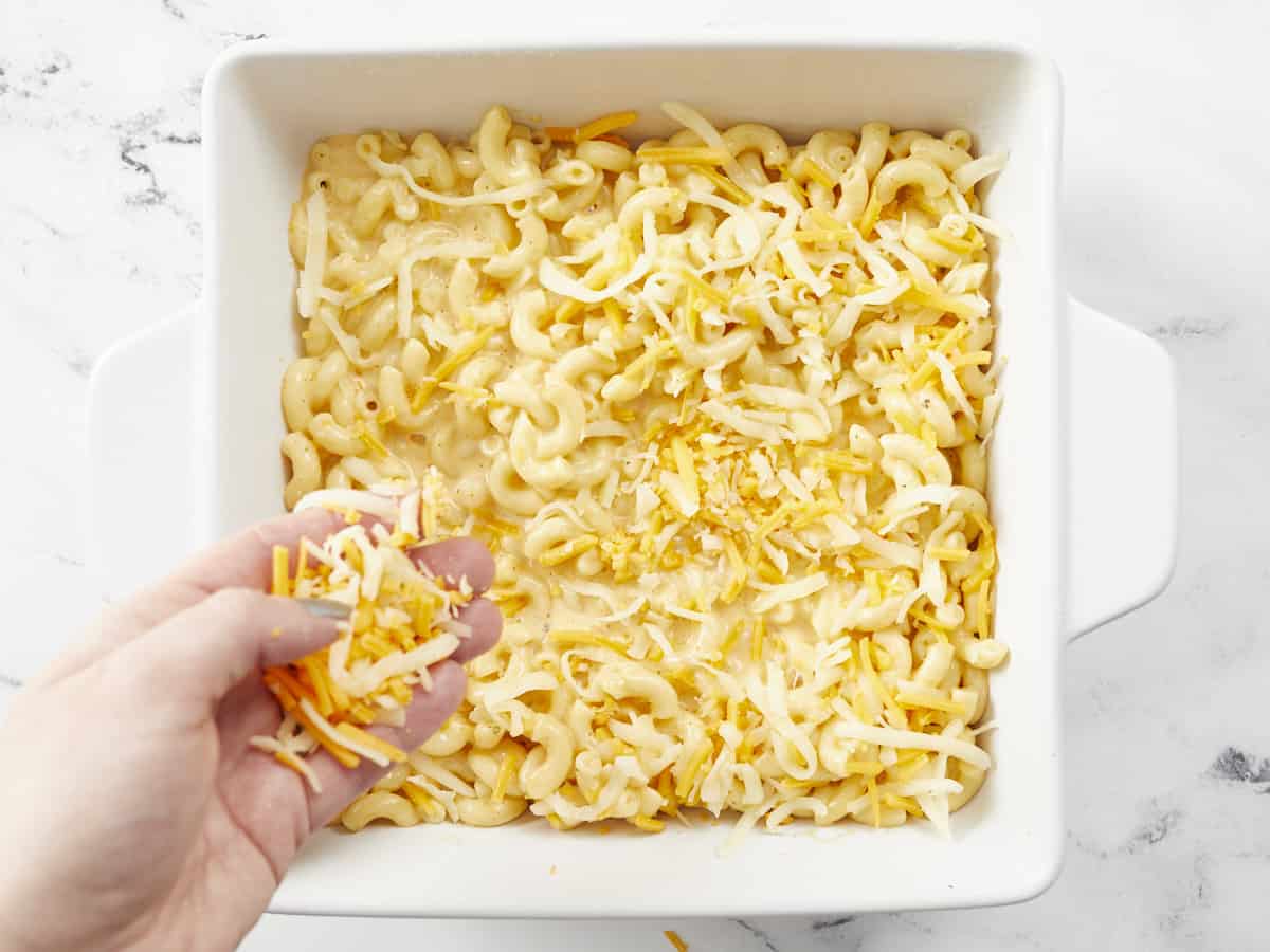 Shredded cheese being layered on top of the mac and cheese in the casserole dish.